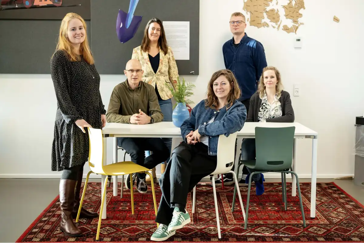 Seks kolleger smiler til kameraet i et kontor, nogle stående og andre siddende omkring et bord, med kunst og et kort på væggen i baggrunden